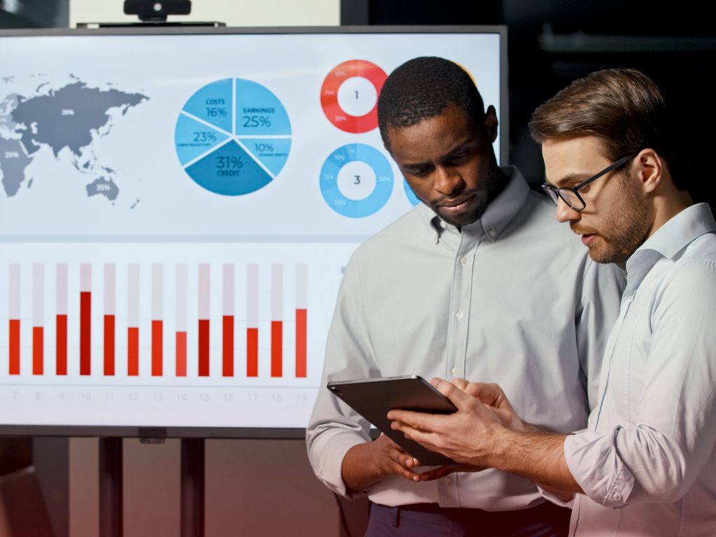 two men standing in a boardroom with an ipad and a screen projecting the suggested sales metrics for their business