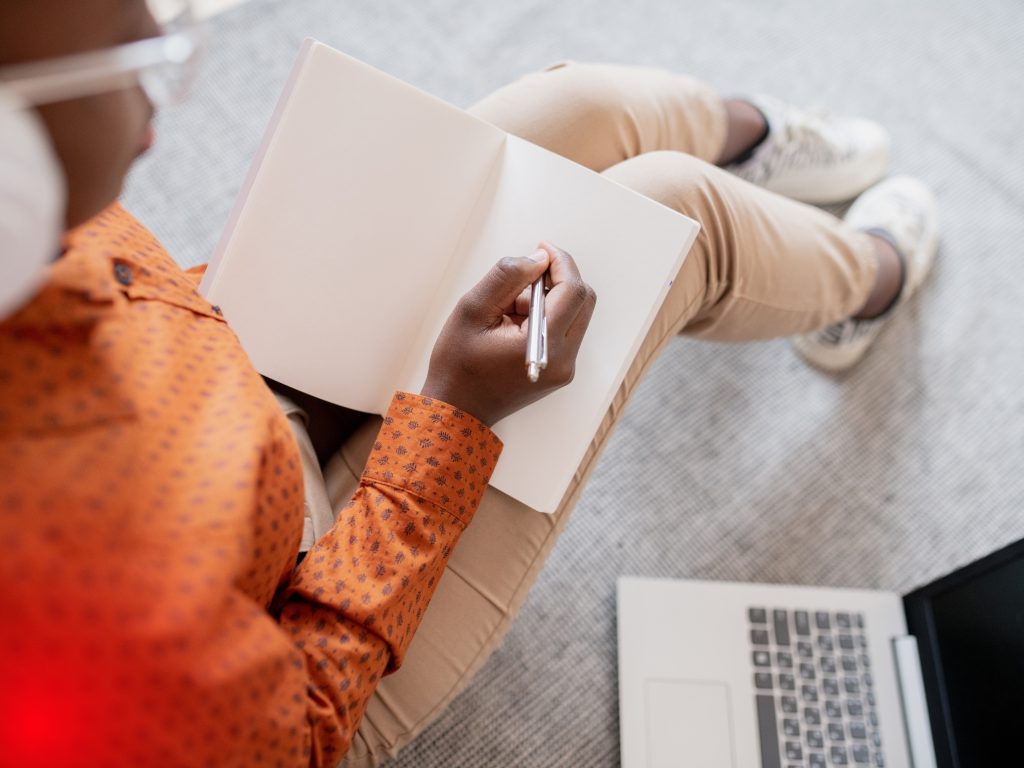 a person sitting on the carpet with bended knees with a notebook on their lap making notes and listening to a podcast about "how to create your own brand story"